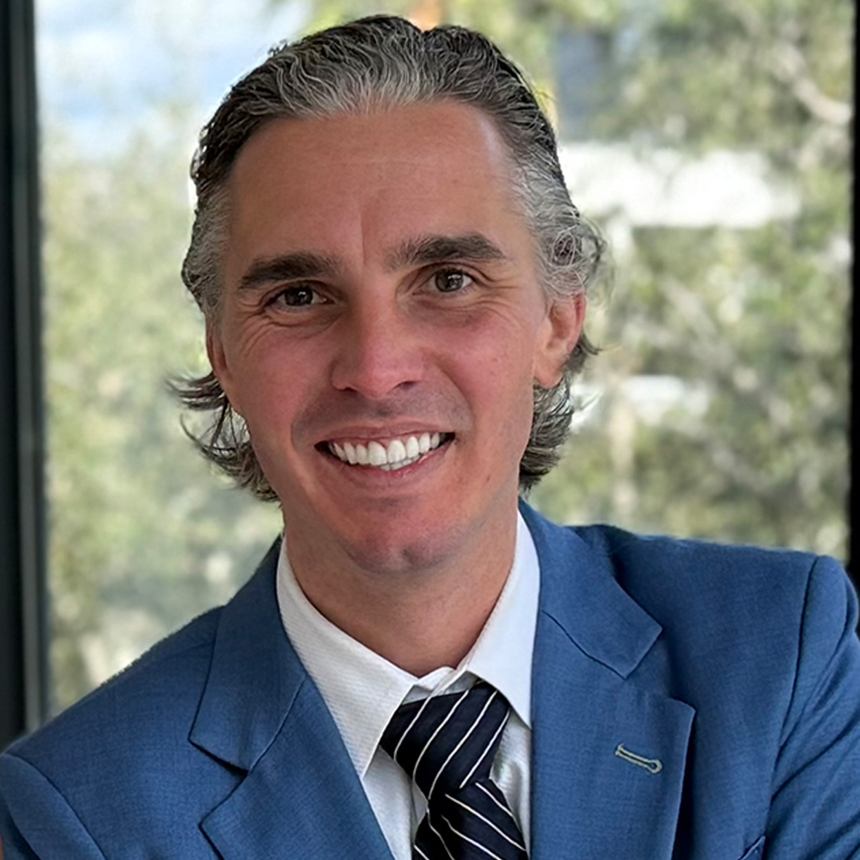 A person with gray hair and a friendly smile is wearing a blue suit, white shirt, and striped tie. They are standing indoors with a blurred, leafy background visible through a window.